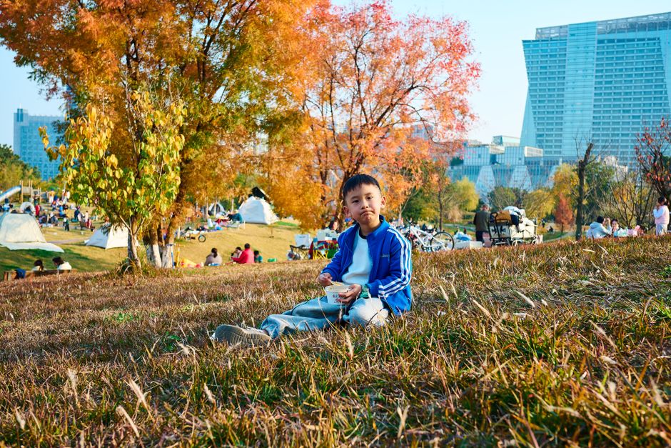 A young boy sitting on grass and eating during a sunny fall day in a busy urban park.