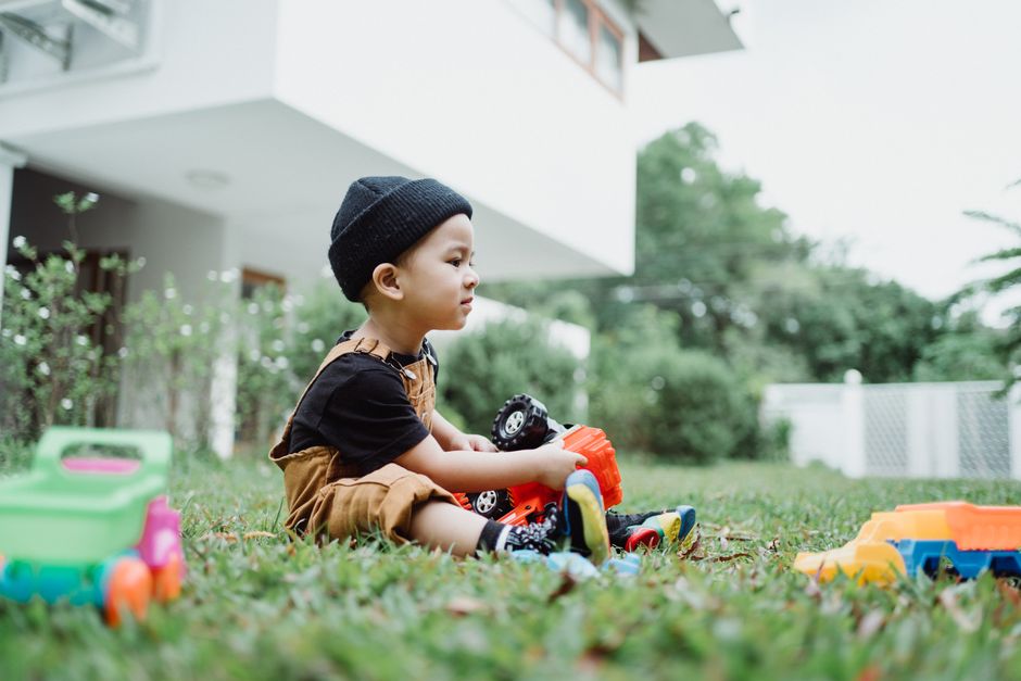 A young boy in overalls and a beanie plays with toy cars on grass outside a modern house.