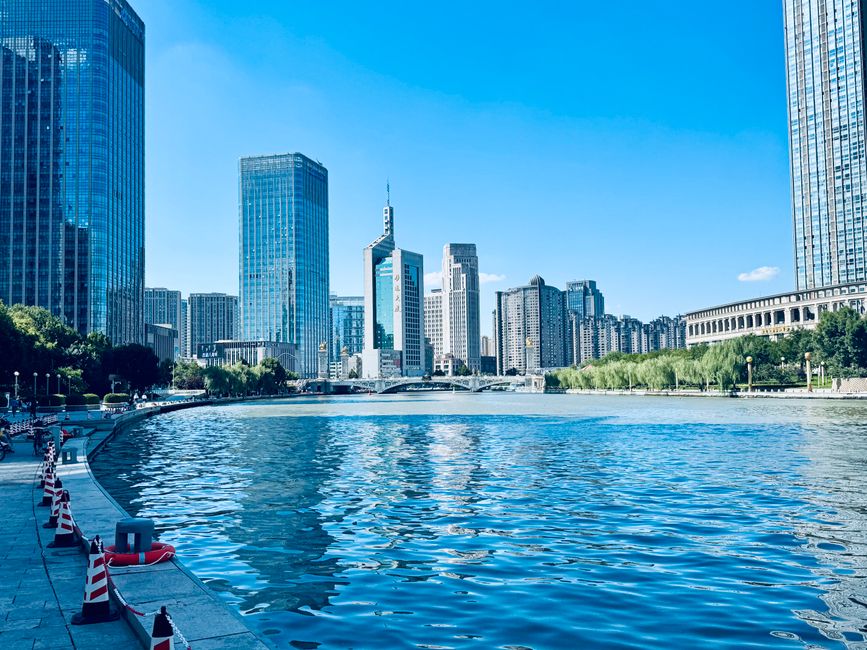 Scenic view of Tianjin's skyscrapers reflecting in a river under the bright blue sky.