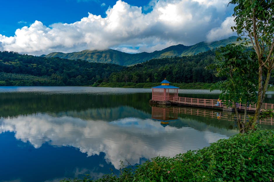 Tranquil lake scene in HirekoLale, India, with a dock, lush greenery, and mountain backdrop under blue skies and fluffy clouds.