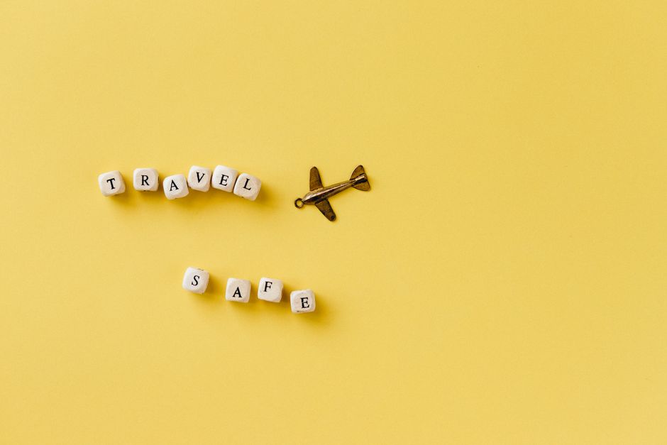 Flat lay of 'Travel Safe' text with airplane pendant on yellow background, evoking travel and safety concepts.
