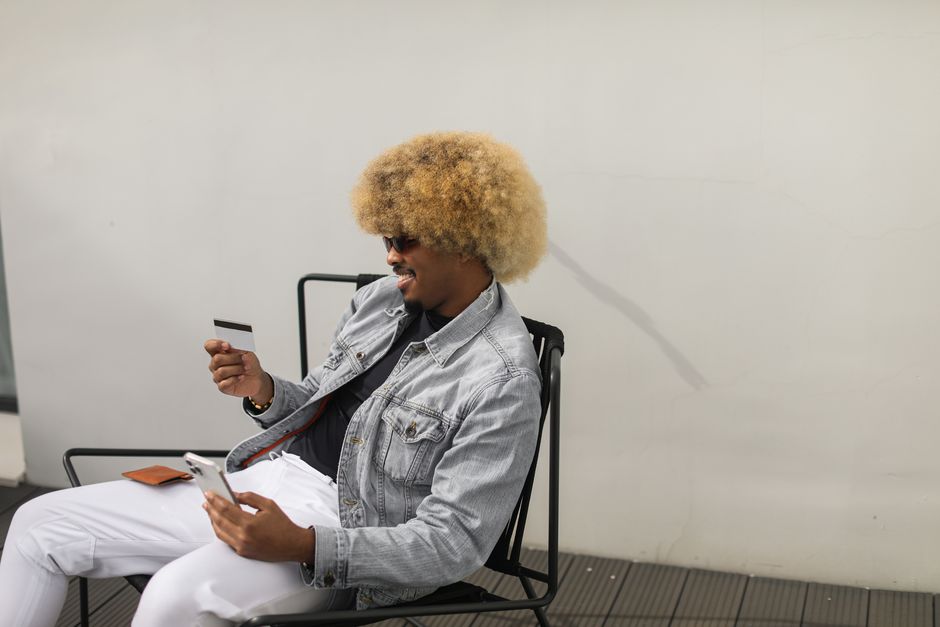 A stylish man with afro hairstyle enjoys online shopping outdoors using his smartphone and credit card.
